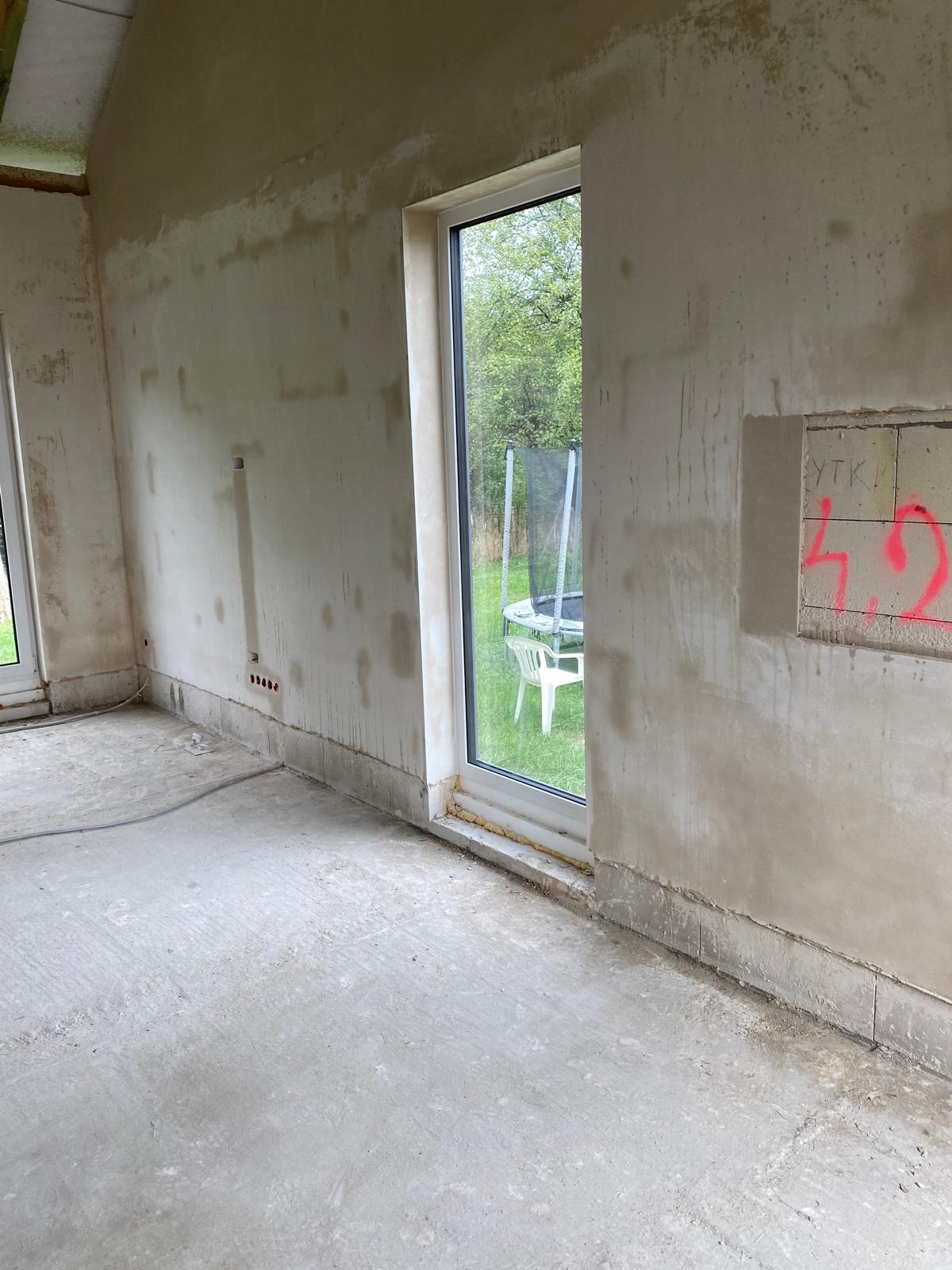 Surowe wnętrze w trakcie prac przygotowawczych: ściany po nałożeniu warstwy szpachli, widoczne ślady po instalacjach, betonowa podłoga, okno z widokiem na ogród z trampoliną i krzesłem, fragment...
