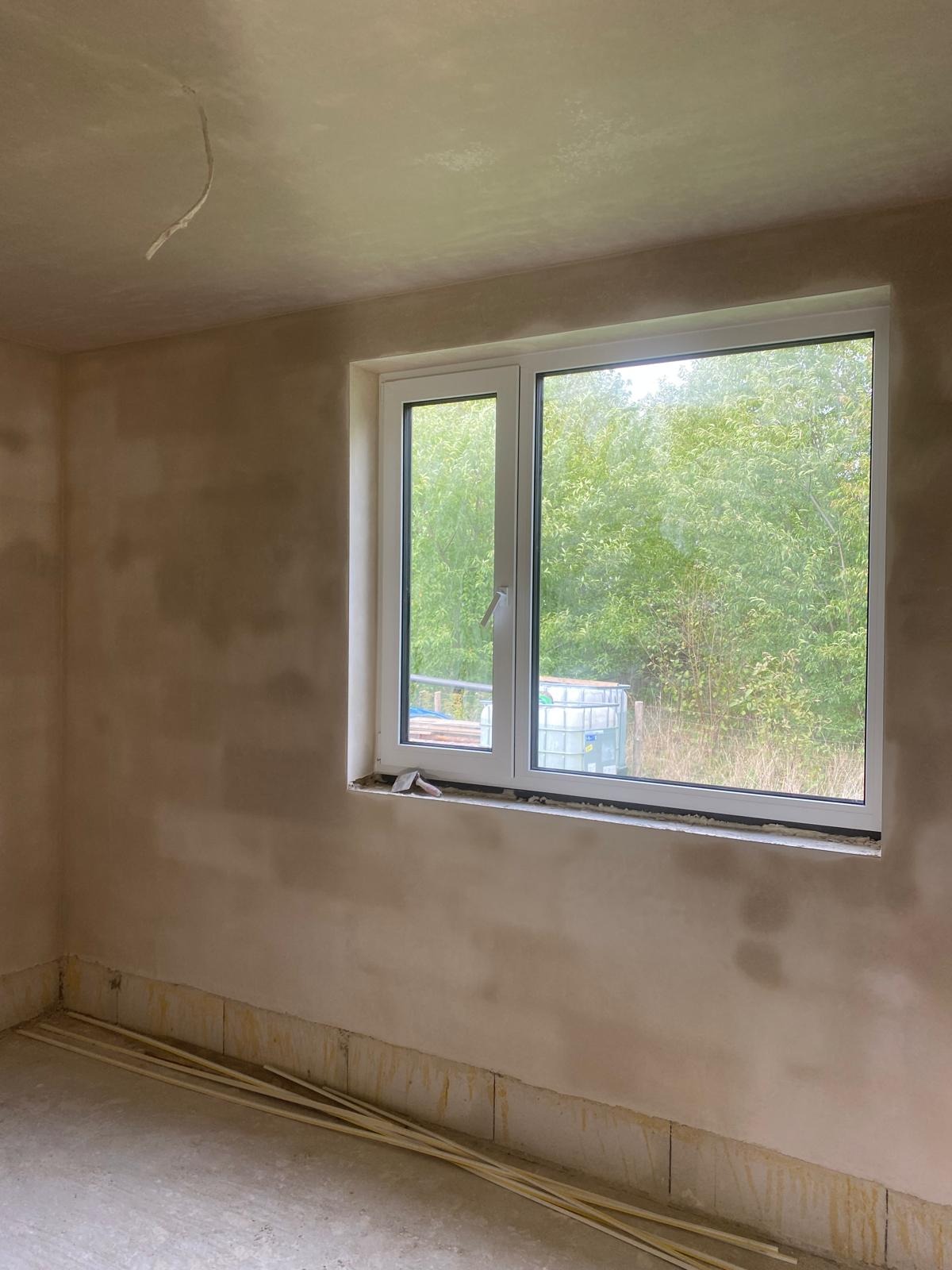 Pokój w trakcie przygotowania do malowania: ściany po wstępnym wyrównaniu, z widocznymi śladami szpachli, białe okno z widokiem na zieleń na zewnątrz, listwy przypodłogowe leżące na podłodze.