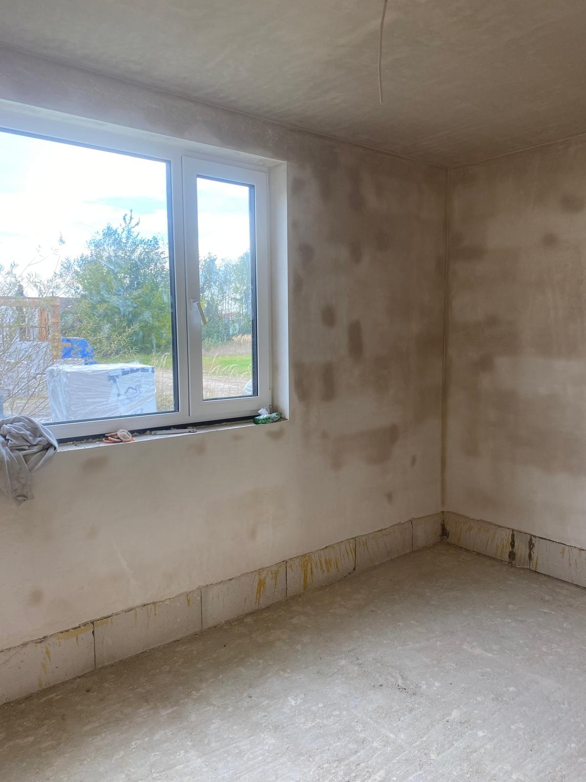 Surowe wnętrze pokoju w Rzeszowie przed malowaniem, z widocznymi śladami po szpachlowaniu na ścianach i podłodze, okno z widokiem na zieleń i materiały budowlane na parapecie.