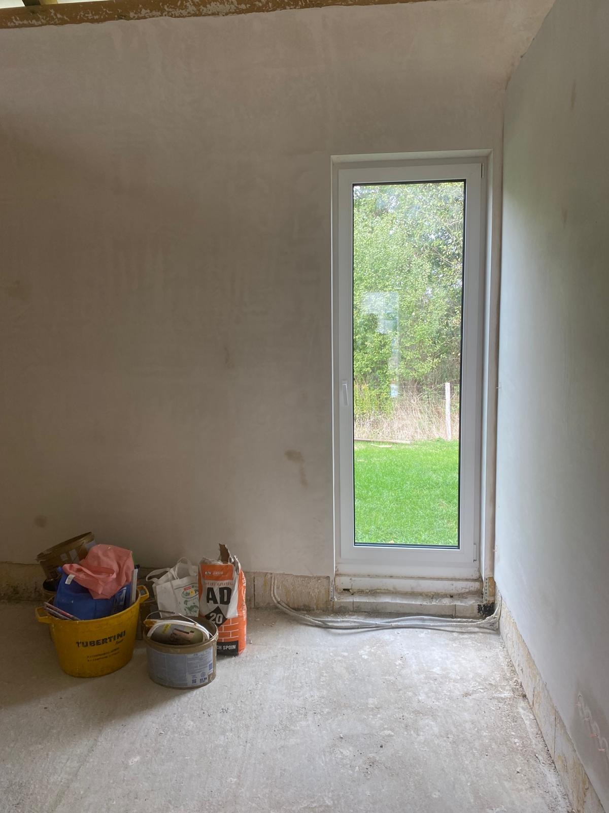 Wnętrze w trakcie prac: ściany po nałożeniu gładzi, widoczny worek zaprawy AD20, żółte wiadro z narzędziami i puszka farby przy ścianie z oknem na ogród.