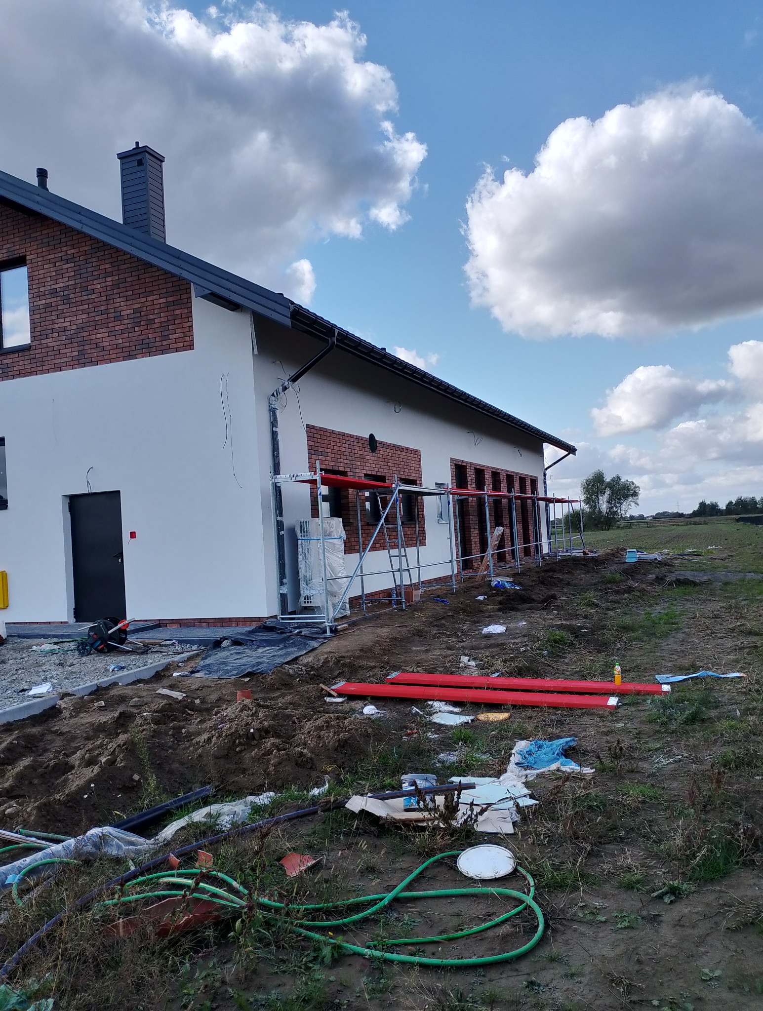 Nowo wybudowany dom z białymi ścianami i elementami z czerwonej cegły, w trakcie prac wykończeniowych, widoczne rusztowania i materiały budowlane wokół.