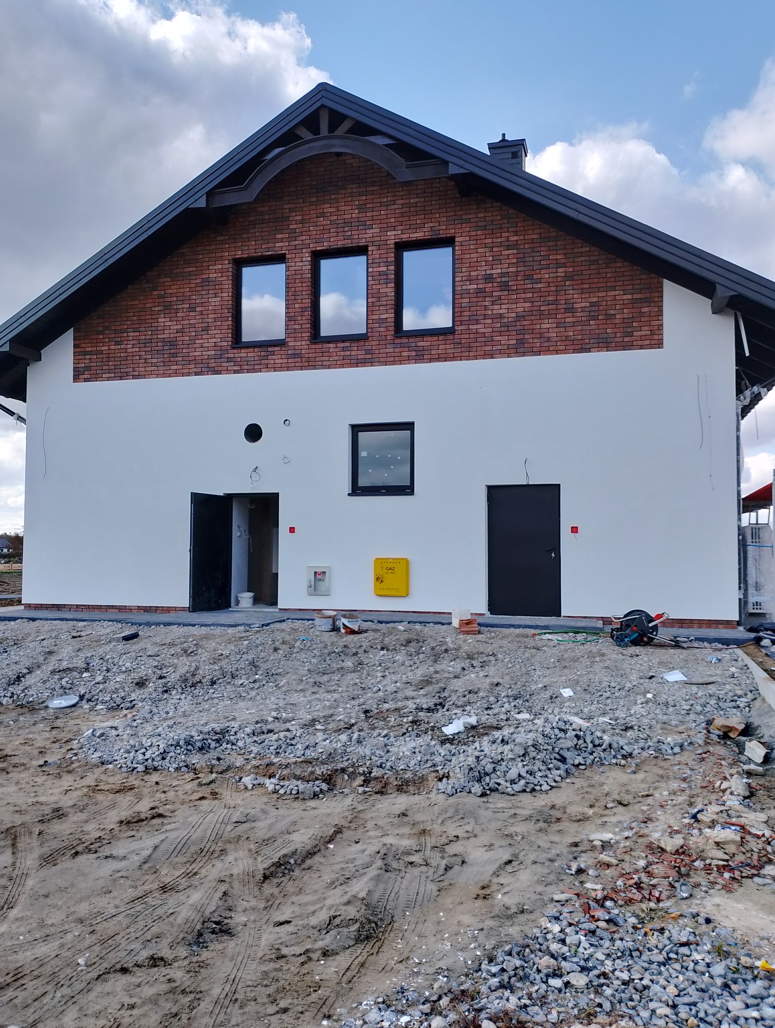 Dwukondygnacyjny dom jednorodzinny z elewacją częściowo wykończoną cegłą klinkierową i pomalowaną na biało, z widocznymi śladami prac wykończeniowych na zewnątrz budynku, w tym gruzem i narzędziami.