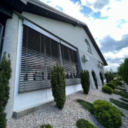 Elewacja domu z zamontowanymi szarymi żaluzjami fasadowymi Z-90, widok z dołu na tle błękitnego nieba z chmurami i ozdobnego ogrodu.