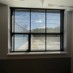 Czarne, poziome żaluzje aluminiowe 50mm z perforowaną taśmą, zamontowane w oknie biurowym z widokiem na budynek i drzewa w tle.