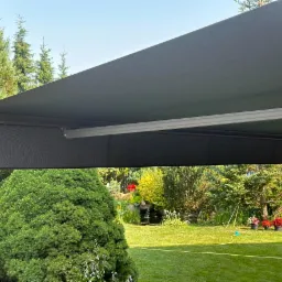 Zewnętrzna markiza tarasowa w kolorze grafitowym, zamontowana nad trawnikiem z dekoracyjnym oczkiem wodnym i rabatami kwiatowymi w tle.