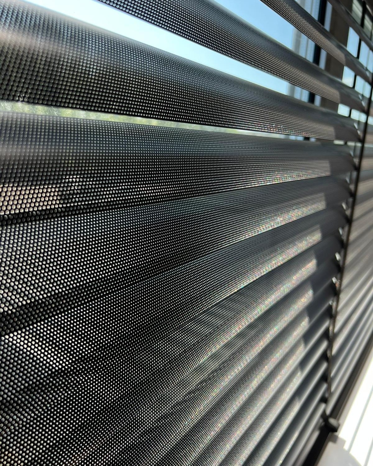 Detaliczne zbliżenie na perforowane, poziome żaluzje aluminiowe w kolorze czarnym matowym, z widocznym światłem słonecznym prześwitującym przez otwory.