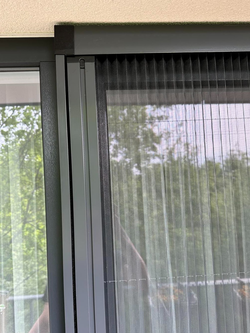 Szczegółowe ujęcie plisowanej siatki na owady w szarej ramie okiennej, widok na zieleń za oknem.