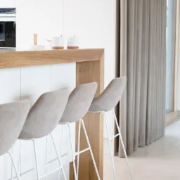 Minimalistyczna kuchnia z białymi szafkami, drewnianym blatem i czterema beżowymi hokerami na białych nogach. Na blacie dwie białe filiżanki z korkowymi podkładkami. W tle beżowe zasłony i okno.