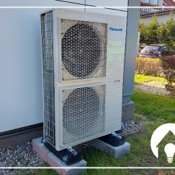 Sollab - Zewnętrzna jednostka pompy ciepła Panasonic zamontowana na betonowych podstawach przy ścianie budynku, z widoczną zielenią i samochodem w tle.