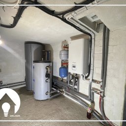 Sollab - Instalacja grzewcza w piwnicy: dwa szare zbiorniki na wodę, biały piec grzewczy wiszący na ścianie, rury instalacyjne rozprowadzone po ścianach i suficie, logo firmy w rogu.
