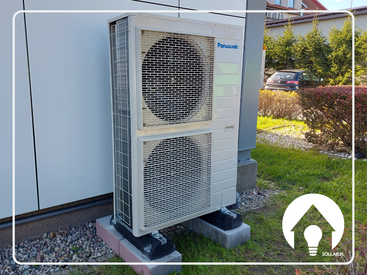 Zewnętrzna jednostka pompy ciepła Panasonic zamontowana na betonowych podstawach przy ścianie budynku, z widoczną zielenią i samochodem w tle.