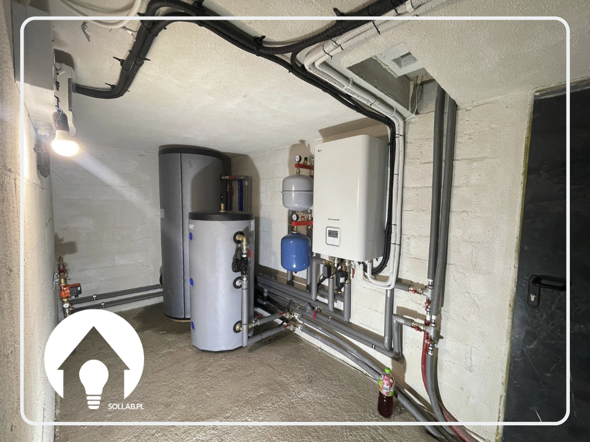 Instalacja grzewcza w piwnicy: dwa szare zbiorniki na wodę, biały piec grzewczy wiszący na ścianie, rury instalacyjne rozprowadzone po ścianach i suficie, logo firmy w rogu.