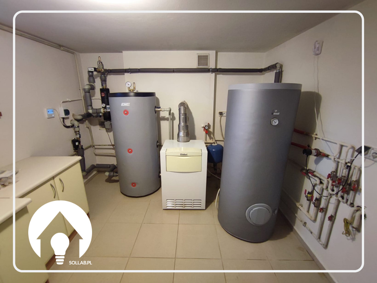 Kotłownia z dwoma szarymi zbiornikami akumulacyjnymi ciepła marki DEMET i Vaillant, połączonymi rurami, z widoczną armaturą i osprzętem na beżowych płytkach podłogowych.