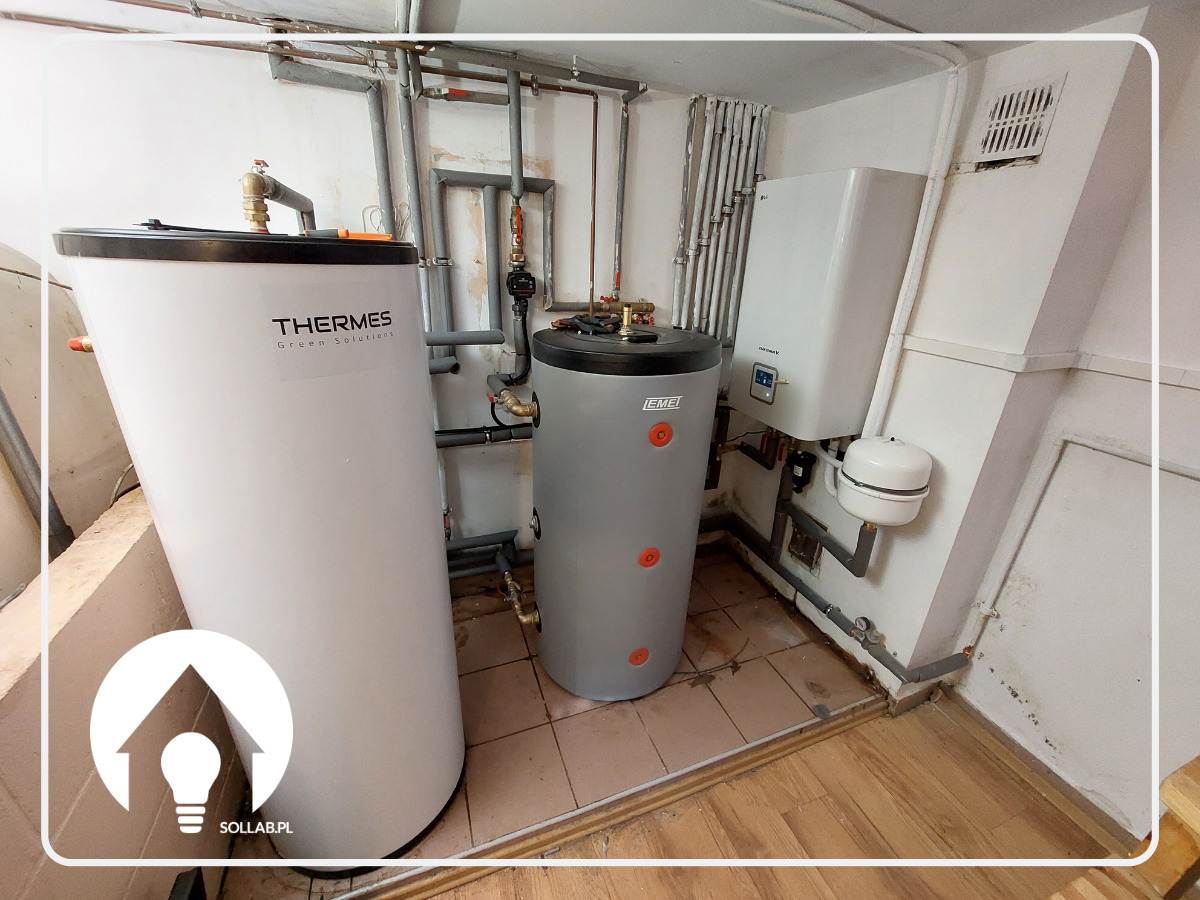 Dwa zbiorniki akumulacyjne ciepła (biały i szary) w kotłowni, obok wiszący na ścianie biały piec gazowy z wyświetlaczem, widoczne rury instalacji grzewczej.