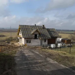 Surowy stan domu z pustaków z przygotowanym szkieletem dachu, widok z polnej drogi, materiały budowlane składowane obok.