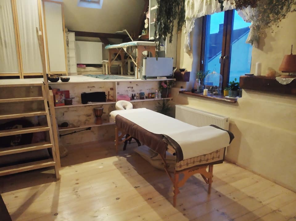 Pokój na poddaszu z drewnianą podłogą, schodami i dwoma stołami do masażu, jeden z białym pokrowcem i brązowym obramowaniem, drugi na podwyższeniu, a także półki z dekoracjami i oknem z widokiem...