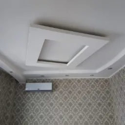 Biały sufit z dekoracyjnym podwieszanym elementem kwadratowym i oświetleniem punktowym, klimatyzator na ścianie pokrytej tapetą w ornamenty.