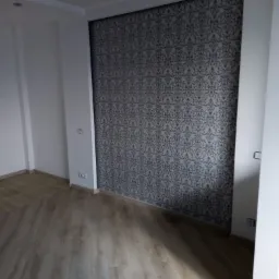 Ściana w pokoju po świeżym tapetowaniu w szaro-białe wzory, z widoczną podłogą z paneli i białymi ścianami bocznymi, z gniazdkami elektrycznymi.