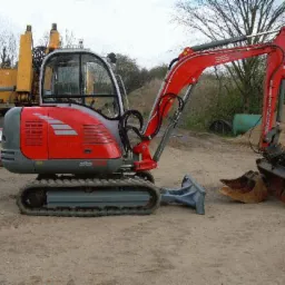 Czerwono-szara mini koparka z łyżką na placu budowy, obok sterta ziemi i inna maszyna w tle. Widoczne gąsienice i kabina operatora.