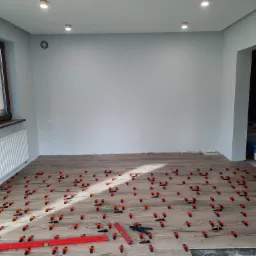 Układanie płytek podłogowych imitujących drewno w jasnym pomieszczeniu, z użyciem klipsów poziomujących i poziomicy, widoczny fragment kominka.