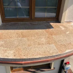 Wykończony taras zewnętrzny z brązowych płyt granitowych, widok z góry, z zaokrąglonym brzegiem i oknem w tle.