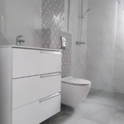 Wykończona łazienka z białą szafką pod umywalkę z trzema szufladami, wiszącą toaletą i kabiną prysznicową ze szklaną ścianką, płytki imitujące marmur na ścianach i podłodze, fragment ściany...