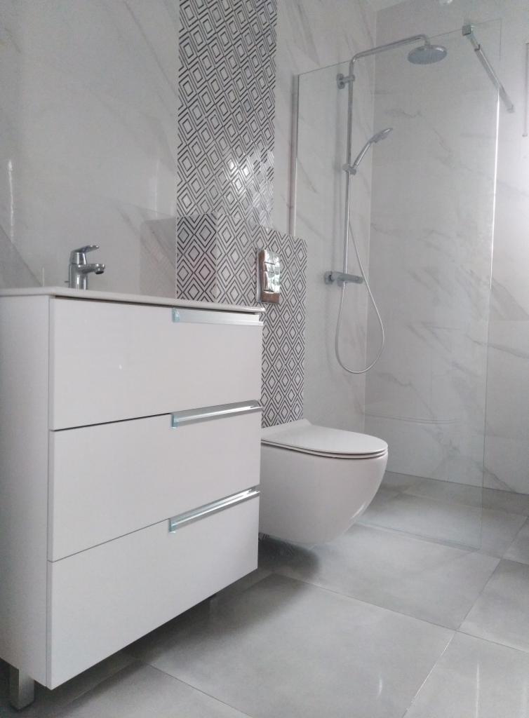 Wykończona łazienka z białą szafką pod umywalkę z trzema szufladami, wiszącą toaletą i kabiną prysznicową ze szklaną ścianką, płytki imitujące marmur na ścianach i podłodze, fragment ściany...