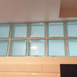 Ścianka z luksferów w łazience, ułożona nad białymi, wypukłymi płytkami, z widocznym fragmentem czarnej zabudowy.