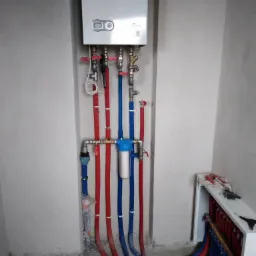 Nowy piec gazowy zamontowany na ścianie z podłączonymi rurami wodnymi w kolorze czerwonym i niebieskim, filtr wody na rurach, widoczne narzędzia hydrauliczne w tle.