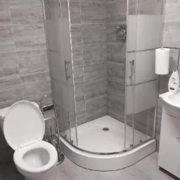 Wykończona łazienka z kabiną prysznicową narożną z częściowo matowymi szybami, toaletą z otwartą klapą i umywalką z białymi szafkami podwieszanymi na tle szarych płytek imitujących drewno.