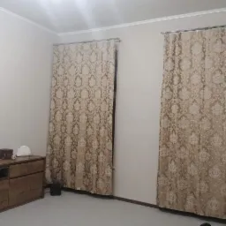 Beżowe, wzorzyste zasłony wiszące na metalowych karniszach w pokoju z drewnianą komodą i jasną podłogą.