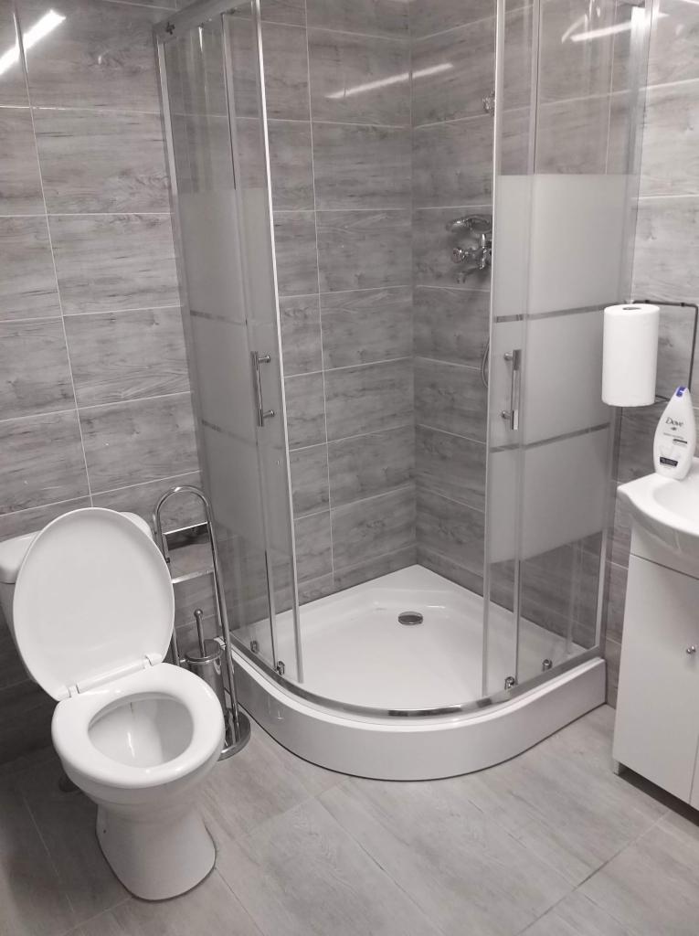 Wykończona łazienka z kabiną prysznicową narożną z częściowo matowymi szybami, toaletą z otwartą klapą i umywalką z białymi szafkami podwieszanymi na tle szarych płytek imitujących drewno.