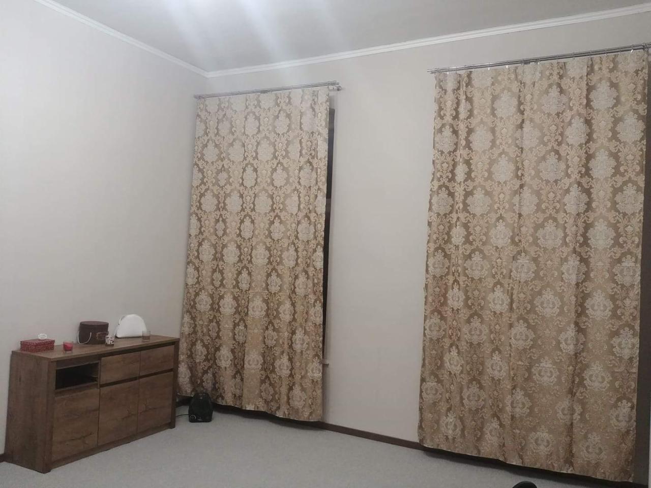 Beżowe, wzorzyste zasłony wiszące na metalowych karniszach w pokoju z drewnianą komodą i jasną podłogą.