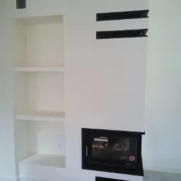 Biała zabudowa kominka z wnękami i półkami, czarny wkład kominkowy z szybą, minimalistyczny design.