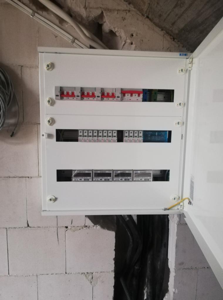 Otwarta biała skrzynka rozdzielcza z zainstalowanymi bezpiecznikami, licznikami i okablowaniem, zamontowana na surowej ścianie z betonu komórkowego w trakcie prac instalacyjnych.