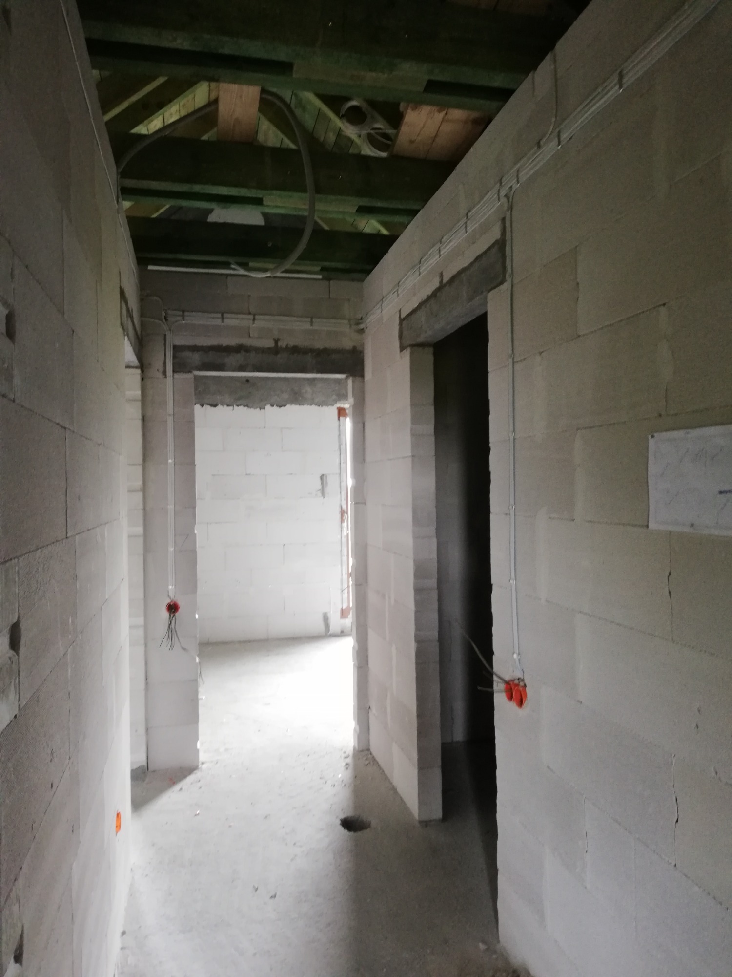 Wnętrze w trakcie budowy z widoczną instalacją elektryczną: kable prowadzone po ścianach z bloczków, puszki instalacyjne z wystającymi przewodami, rury ochronne pod sufitem.
