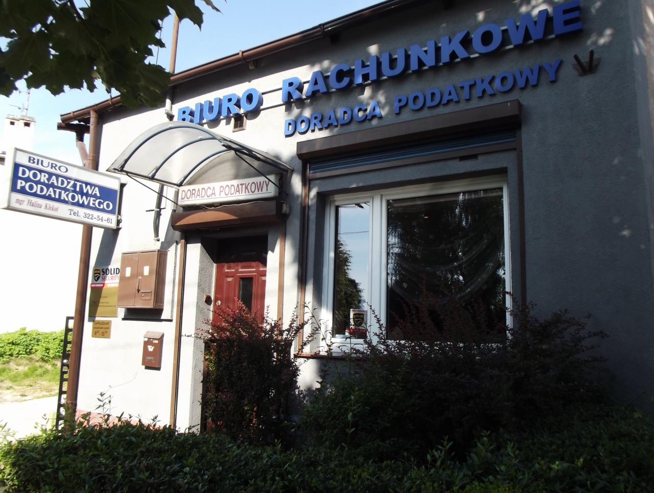 Fasada budynku biura rachunkowego z niebieskim napisem 'BIURO RACHUNKOWE DORADCA PODATKOWY', tabliczka informacyjna 'BIURO DORADZTWA PODATKOWEGO' oraz okno z roletą.