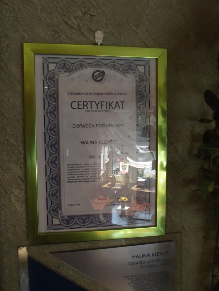 Certyfikat doradcy podatkowego Haliny Klekot w złotej ramie, wiszący na ścianie z kamienną fakturą, pod nim tabliczka informacyjna z danymi doradcy.