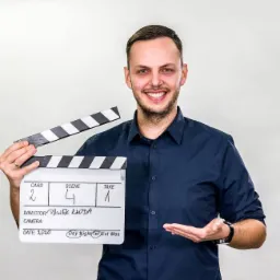 Uśmiechnięty mężczyzna w granatowej koszuli trzyma klaps filmowy z wpisanymi danymi produkcji: Card 2, Scene 4, Take 1, Director Paweł Kłoda, Data 2020.