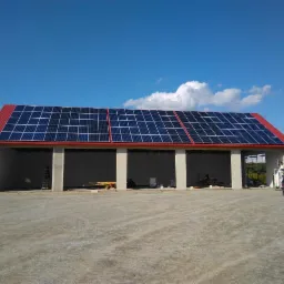 Budynek z czerwonym dachem i panelami słonecznymi w trakcie instalacji, widoczne rusztowania i samochód dostawczy z otwartymi drzwiami bagażnika, błękitne niebo z chmurami w tle.