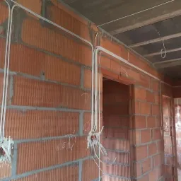 Wewnętrzne ściany z cegły dziurawki w trakcie budowy, z poprowadzonymi białymi przewodami elektrycznymi w peszlach, widoczne puszki instalacyjne z wystającymi kablami.