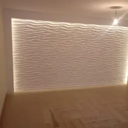 Ściana z dekoracyjnymi panelami 3D o falistym wzorze, podświetlona od góry i dołu, z widoczną podłogą z płytek imitujących drewno i butelką kleju w rogu.