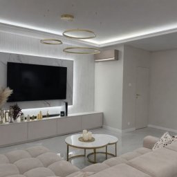 Usługi budowlane Cezary Piaskowski - Elegancki salon w jasnych barwach z telewizorem na ścianie z marmurowym panelem i dekoracyjnym oświetleniem. Widoczny beżowy narożnik i stolik kawowy.