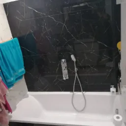 Wykończona łazienka z wanną obudowaną czarnymi płytkami imitującymi marmur z jasnymi żyłami, chromowana bateria wannowa z prysznicem, turkusowy ręcznik po lewej stronie.