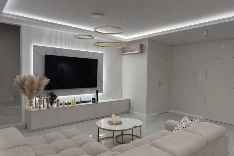 Elegancki salon w jasnych barwach z telewizorem na ścianie z marmurowym panelem i dekoracyjnym oświetleniem. Widoczny beżowy narożnik i stolik kawowy.