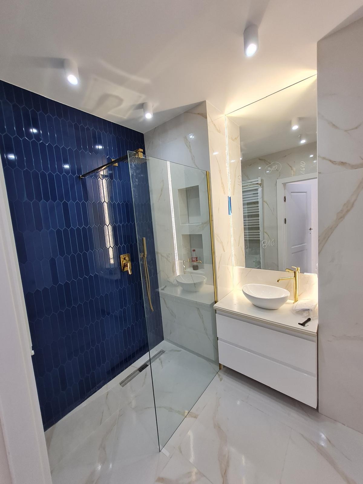 Nowoczesna łazienka po remoncie: kobaltowa ściana z podłużnych płytek, kabina prysznicowa ze złotymi elementami, biała umywalka nablatowa i marmurowe płytki podłogowe.