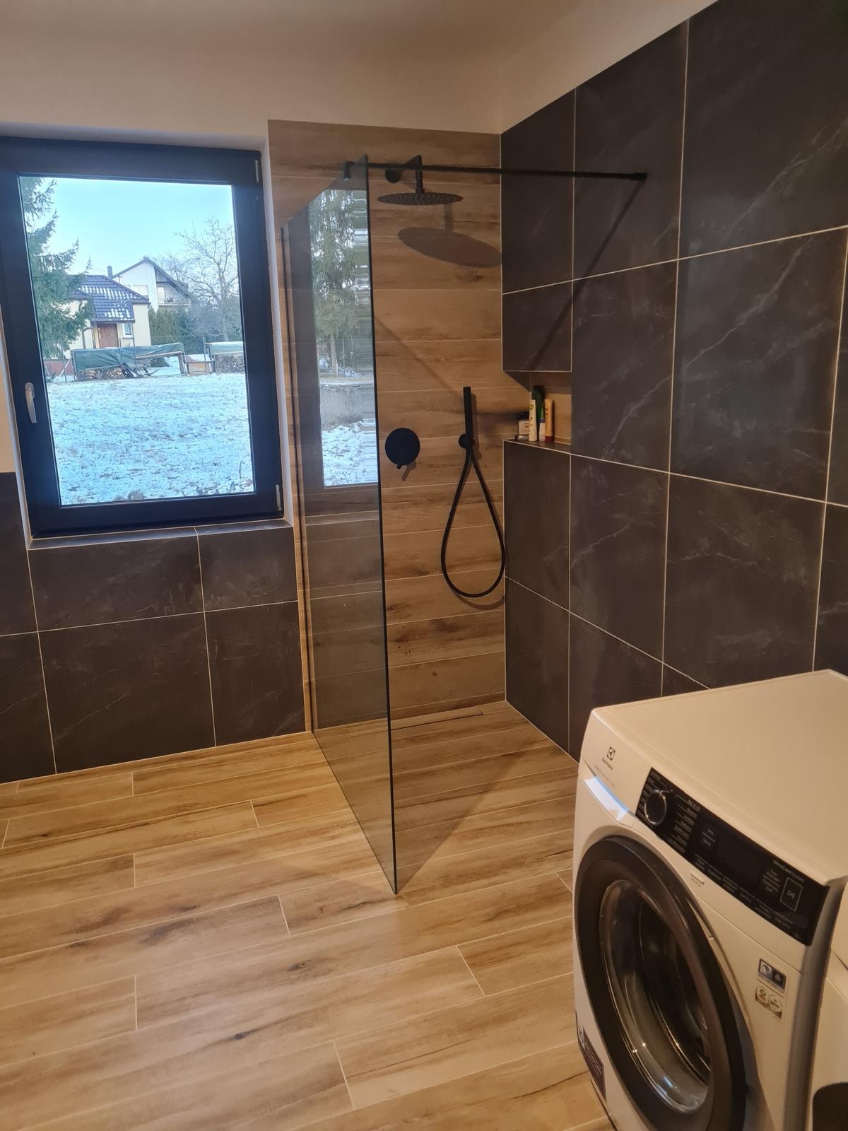 Nowoczesna łazienka z kabiną prysznicową typu walk-in, wykończona płytkami imitującymi drewno i ciemnym marmurem, z widoczną pralką i oknem na zimowy krajobraz.