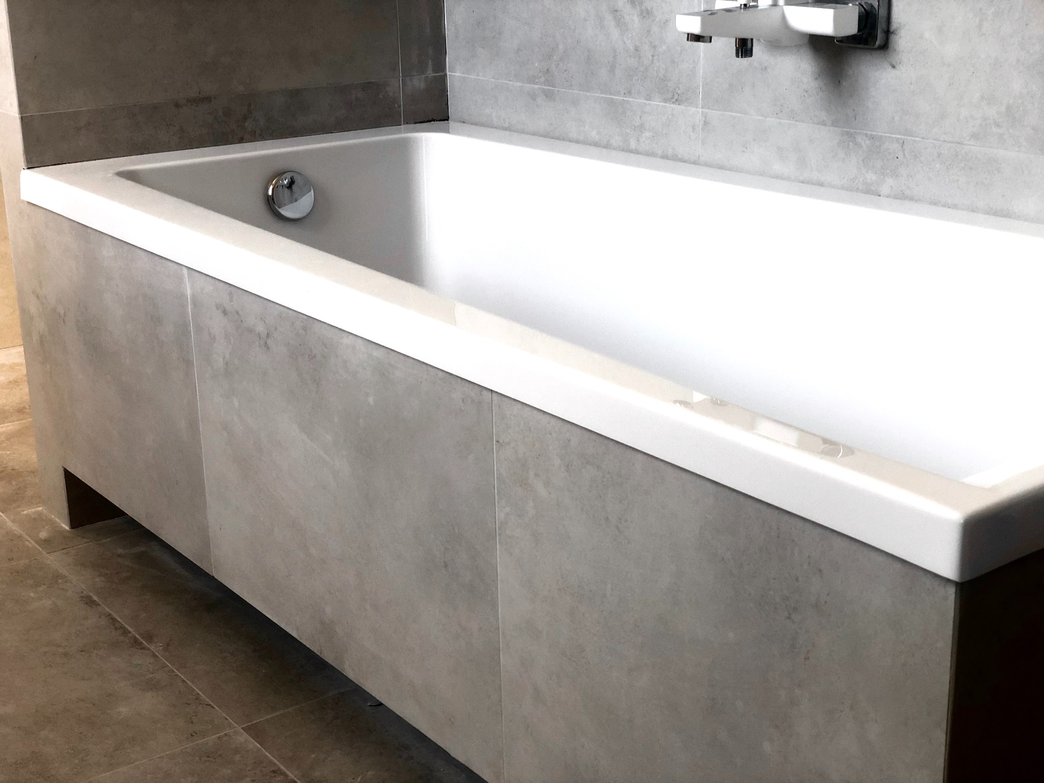 Nowoczesna, biała wanna obudowana szarymi płytami imitującymi beton, z chromowaną armaturą na ścianie z płytek w odcieniach szarości. Minimalistyczny design łazienki.