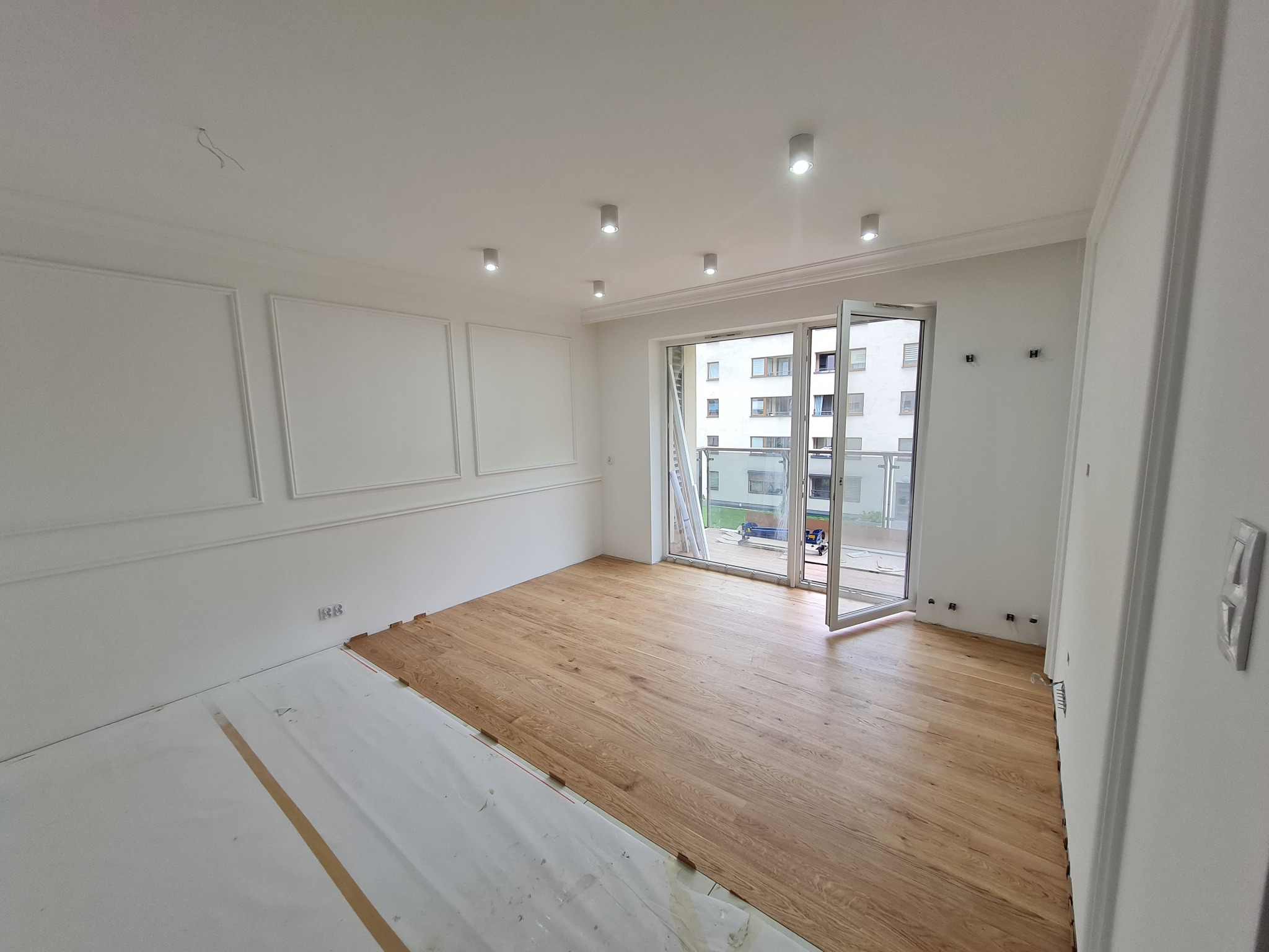 Jasne panele podłogowe w trakcie układania w przestronnym pokoju z białymi ścianami i sztukaterią. Część podłogi zabezpieczona folią ochronną.