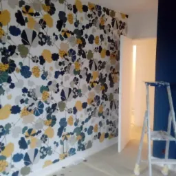 Ściana w pokoju z tapetą w kwiaty w odcieniach granatu, żółci i szarości, zabezpieczona folią podłoga, drabina malarska w rogu, widok na białe drzwi i fragment ściany w kolorze granatowym.